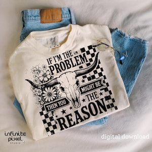 May include: Cream-colored t-shirt with a graphic featuring a bull skull, flowers, and the text "IF I'M THE PROBLEM THEN YOU MIGHT BE THE REASON." The shirt is paired with light blue jeans and round glasses.
