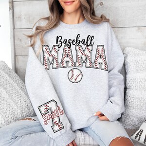Pu&ograve; includere: Felpa grigia con il testo "Baseball Mama" in nero con una grafica di baseball. La manica ha una grafica di baseball con il testo "Smith" in rosso.
