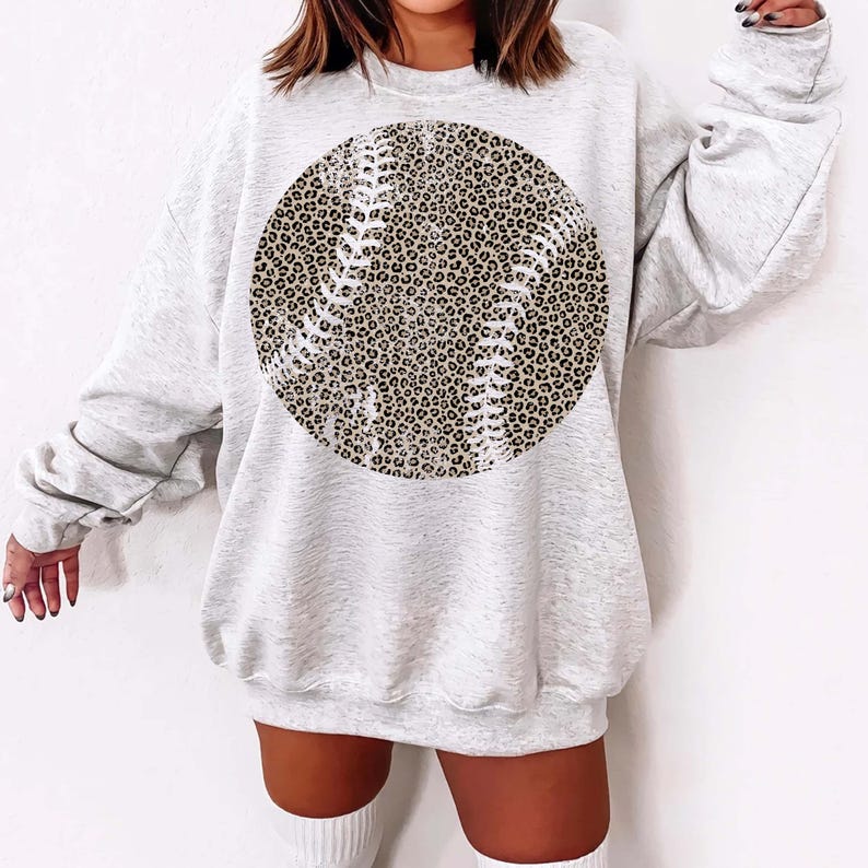 May include: A light gray sweatshirt featuring a large circular baseball design. The baseball graphic has a leopard print pattern with white stitching details. The sweatshirt is long-sleeved and has a relaxed fit.
