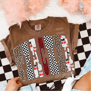 May include: A brown t-shirt with the word "TEACHER" in a bold, decorative font. The letters feature a mix of leopard print, checkered patterns, and cherry designs. The shirt has a vintage, worn look.