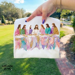 May include: White embroidered t-shirt featuring a colorful depiction of the Last Supper. The design includes figures in various colored robes seated at a table with a detailed, textured tablecloth. The shirt is held up by a hand. The logo "infinite pixel studio" is at the bottom.