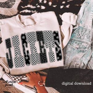 May include: Cream-colored sweatshirt with the word "JACKETS" in a bold, stylized font. The letters feature a mix of patterns, including checkered, leopard print, and star designs. A denim jacket and checkered shoes are also visible.