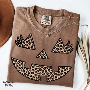 May include: A brown t-shirt with a jack-o'-lantern face design. The face features leopard print accents for the eyes, nose, and mouth. A silver necklace with a pendant is draped across the shirt. The shirt is folded and has a Comfort Colors tag.