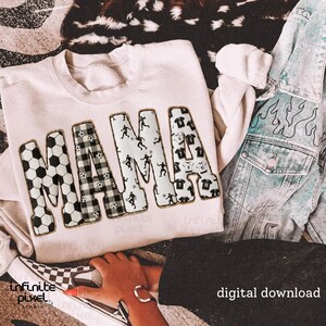 May include: Cream-colored sweatshirt with the word "MAMA" in large, decorative letters. The letters are patterned with soccer balls, black and white plaid, and soccer player silhouettes. A denim jacket with flame designs is visible.