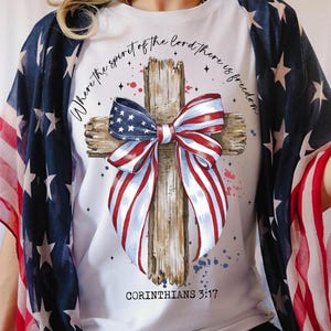 K&ouml;nnte beinhalten: Wei&szlig;es T-Shirt mit einem Holzkreuz und einer rot-wei&szlig;-blauen Schleife. Die Schleife ist aus Stoff mit dem Muster der amerikanischen Flagge. Der Text "Where the spirit of the Lord there is freedom" ist um das Kreuz herum geschrieben. Der Text "Corinthians 3:17" ist unter dem Kreuz geschrieben.