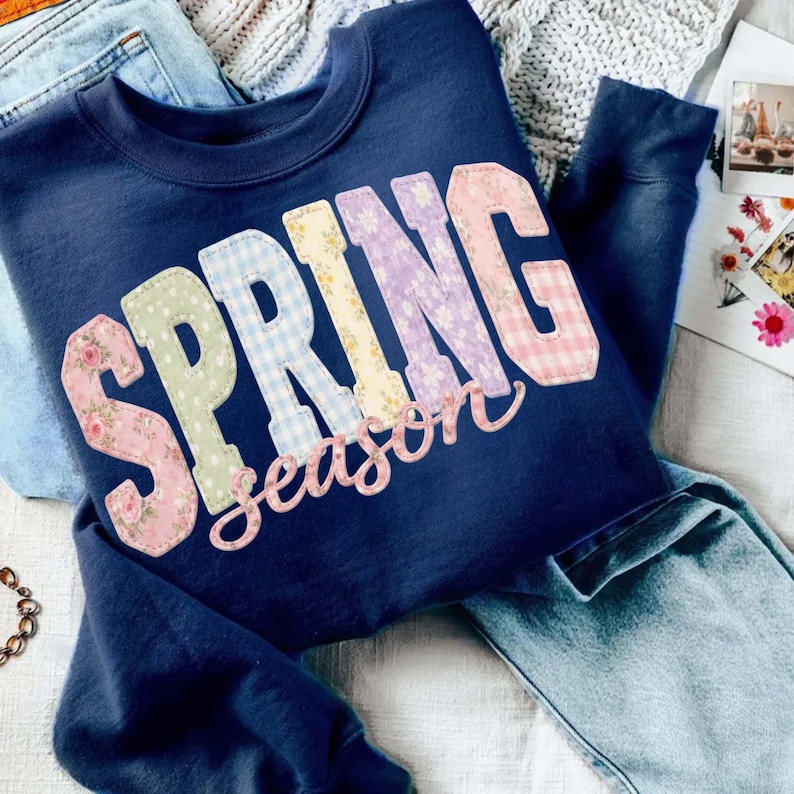 May include: Navy blue sweatshirt with the words "SPRING season" in a patchwork design. The letters are in floral, gingham, and solid patterns. The sweatshirt is folded on top of blue jeans.