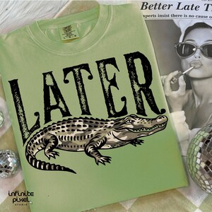 Peut inclure: T-shirt vert clair avec le mot "LATER" en grosses lettres noires et une illustration d&eacute;taill&eacute;e d'un alligator. Le t-shirt a un col rond et des manches courtes. Un magazine et une boule disco sont &eacute;galement visibles.