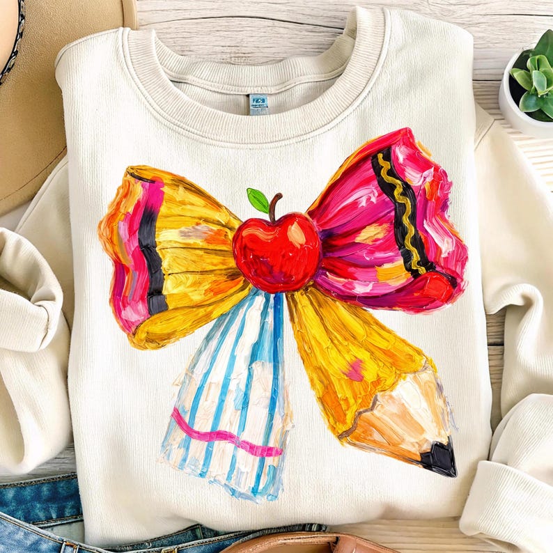 May include: Cream-colored sweatshirt featuring a colorful bow design. The bow is composed of a red apple, yellow pencil, and blue and white striped ribbon. The bow has a painted, artistic style.