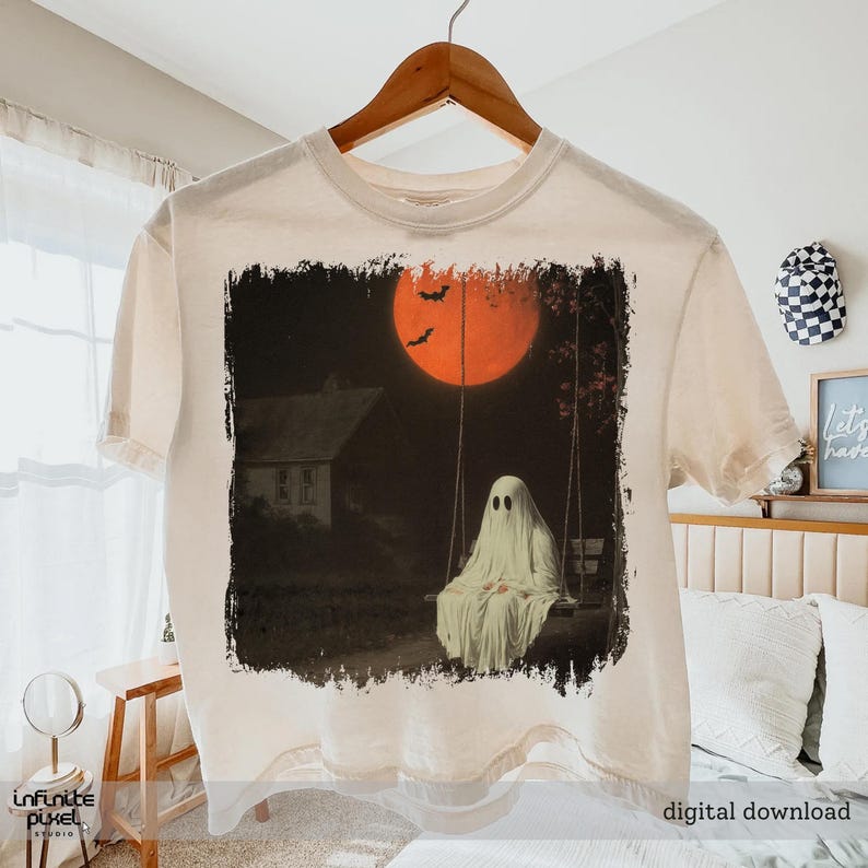 May include: Cream-colored t-shirt featuring a spooky Halloween design. The graphic depicts a ghost on a swing set under a large orange moon with bats. The words "digital download" are printed at the bottom.