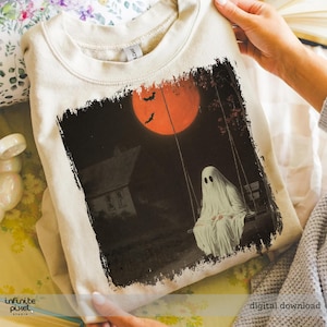 May include: A beige sweatshirt featuring a Halloween-themed graphic. The design depicts a ghost on a swing set under a large orange moon, with bats flying in the sky and a house in the background. The words "digital download" are visible.