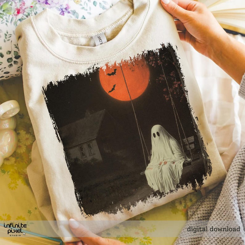 May include: A beige sweatshirt featuring a Halloween-themed graphic. The design depicts a ghost on a swing set under a large orange moon, with bats flying in the sky and a house in the background. The words "digital download" are visible.