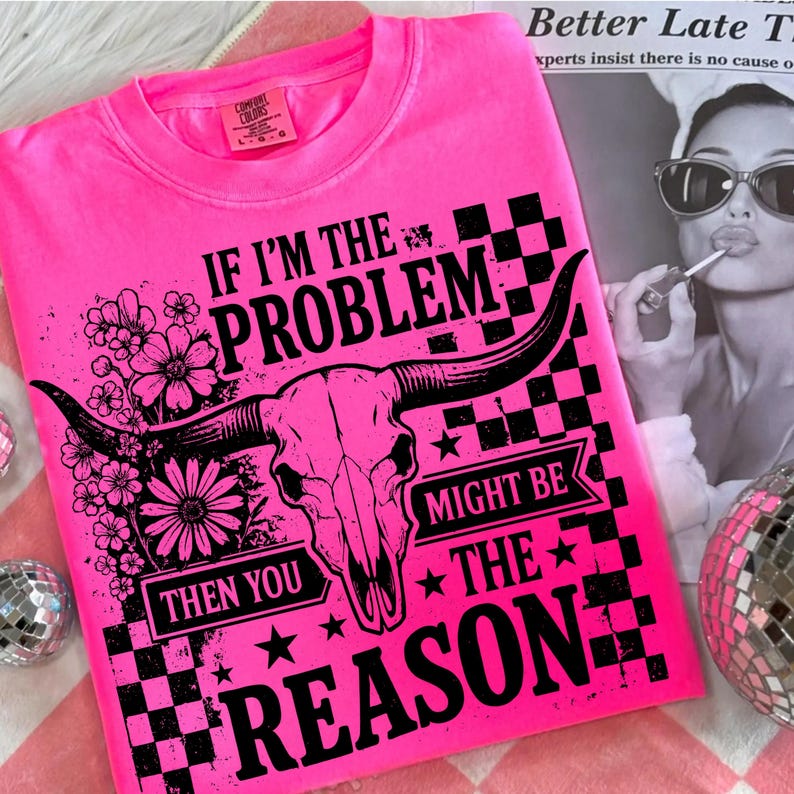 May include: Bright pink sweatshirt with the text "IF I'M THE PROBLEM THEN YOU MIGHT BE THE REASON" and a bull skull graphic. The design includes flowers, stars, and a checkered pattern. The shirt is a Comfort Colors brand.