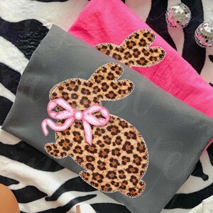 May include: Two decorative pillows, one grey and one pink, with a leopard print bunny design. The bunny has a pink bow. The pillows are on a zebra-striped surface with disco balls.