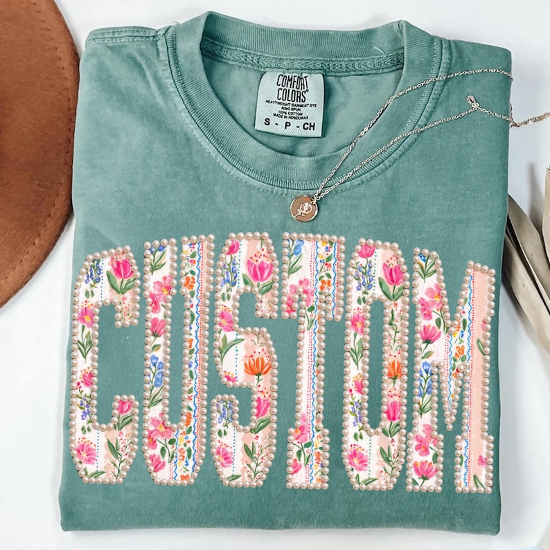 Puede incluir: Una camiseta verde salvia con la palabra "CUSTOM" en letras grandes y florales. Las letras est&aacute;n delineadas con peque&ntilde;as cuentas perladas. Un collar plateado con un peque&ntilde;o colgante descansa sobre la camiseta.