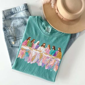 May include: A teal-colored t-shirt featuring an embroidered image of the Last Supper. The shirt is paired with light-wash denim jeans, a gold necklace, and a tan fedora hat. The image is set against a white background.