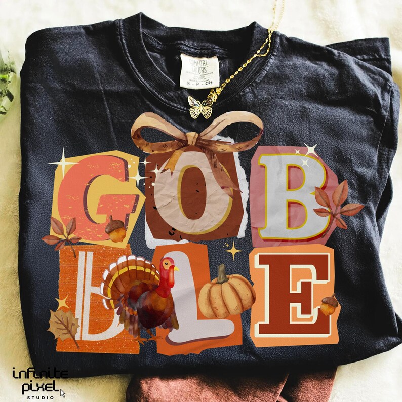 May include: Black t-shirt with a colorful Thanksgiving-themed graphic. The word "GOBBLE" is spelled out in large, block letters with autumn-themed illustrations. A gold butterfly necklace is draped on the shirt.