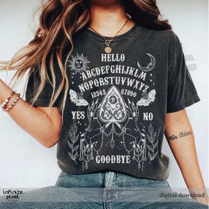 May include: Charcoal grey t-shirt featuring a white Ouija board graphic. The design includes the alphabet, numbers, and the words "Hello," "Yes," "No," and "Goodbye." The shirt also has sun, moon, and butterfly illustrations.
