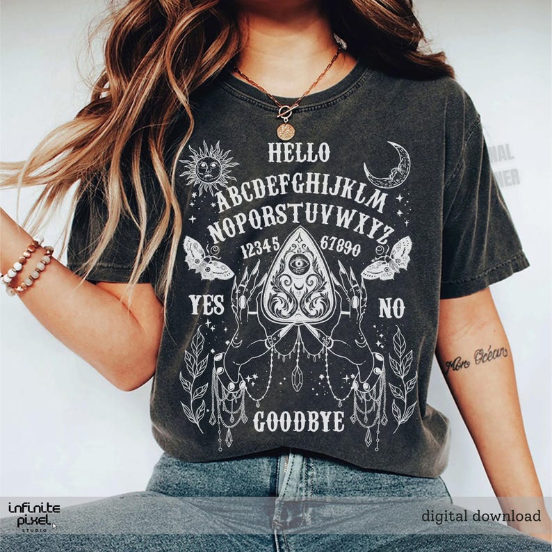May include: Charcoal grey t-shirt featuring a white Ouija board graphic. The design includes the alphabet, numbers, and the words "Hello," "Yes," "No," and "Goodbye." The shirt also has sun, moon, and butterfly illustrations.