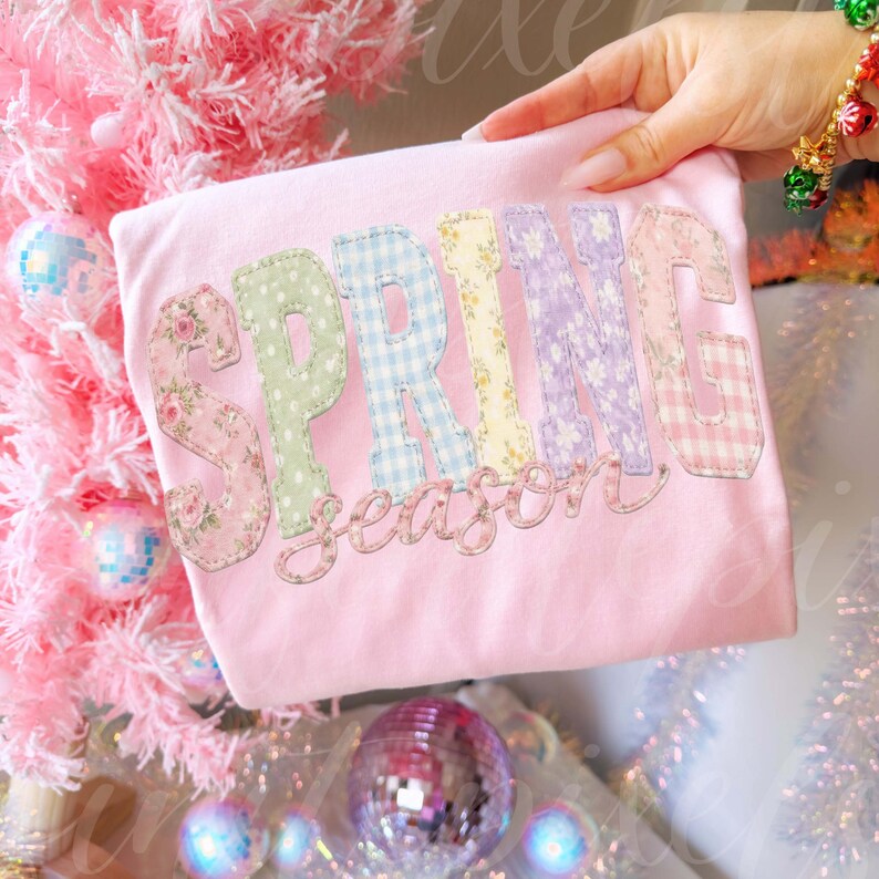 May include: A light pink t-shirt with the word "SPRING" in patchwork letters. The letters are decorated with floral, polka dot, and gingham patterns. The word "season" is written in a cursive font below the word "SPRING".