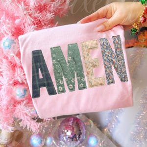 May include: A light pink t-shirt with the word "AMEN" appliqued across the front. The letters are made of various patterned fabrics, including plaid, floral, and gingham. The shirt is held up by a hand, with a pink Christmas tree in the background.