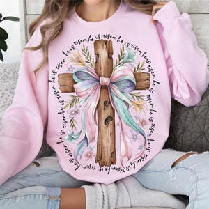 May include: A pink sweatshirt with a white cross and a pink bow. The cross is surrounded by flowers and the text "He is risen. He is risen. He is risen. He is risen. He is risen. He is risen. He is risen. He is risen." is printed around the cross.