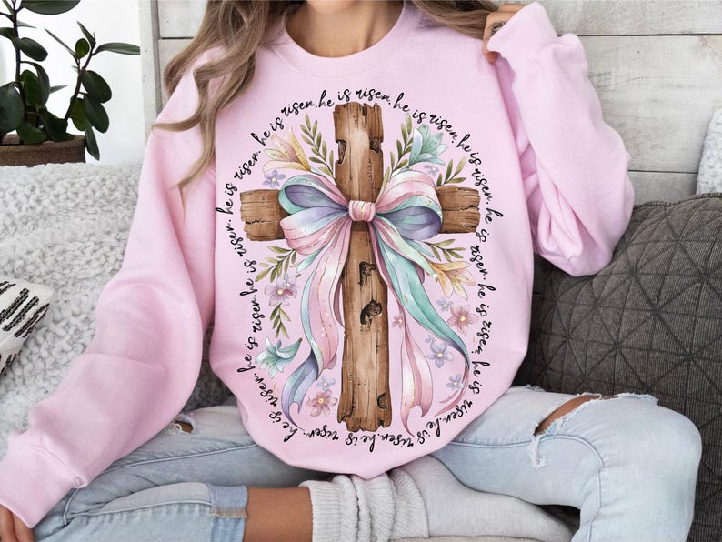 May include: A pink sweatshirt with a white cross and a pink bow. The cross is surrounded by flowers and the text "He is risen. He is risen. He is risen. He is risen. He is risen. He is risen. He is risen. He is risen." is printed around the cross.