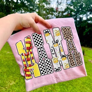 May include: Pink t-shirt with the word "MAMA" in a bold, colorful design. Each letter features a different pattern: yellow with red stripes, black and white checkered, softball and bow, and leopard print. The shirt is held up by a hand.