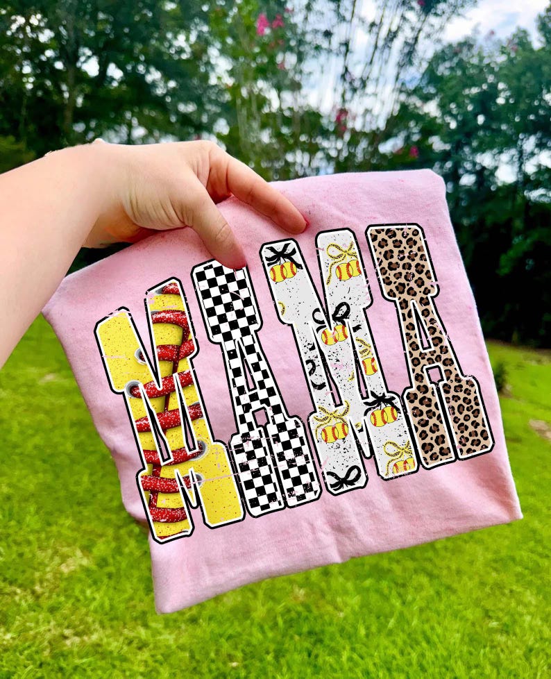 May include: Pink t-shirt with the word "MAMA" in a bold, colorful design. Each letter features a different pattern: yellow with red stripes, black and white checkered, softball and bow, and leopard print. The shirt is held up by a hand.