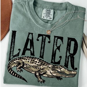 Peut inclure: T-shirt vert sauge avec le mot "LATER" en grandes lettres noires et une illustration d'alligator. Le t-shirt a un col rond et des manches courtes. Un collier argent&eacute; avec un petit pendentif est drap&eacute; sur le t-shirt.