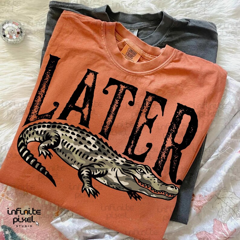 Peut inclure: Un t-shirt orange avec le mot "LATER" en grosses lettres noires et une illustration d&eacute;taill&eacute;e d'un alligator. Un t-shirt gris est visible derri&egrave;re. Les t-shirts sont probablement en coton.