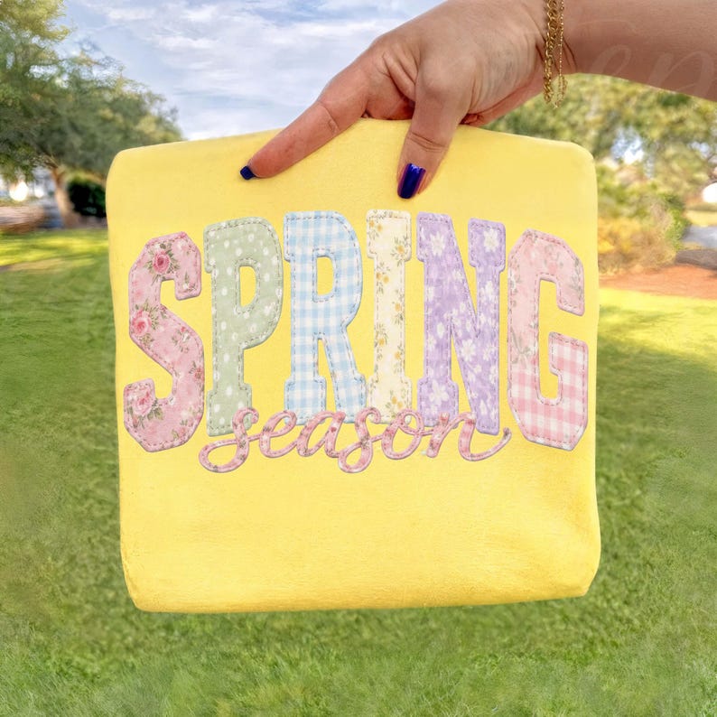 May include: A yellow rectangular pillow with the words "SPRING season" appliqued on the front. The letters are in various pastel patterns, including floral, gingham, and polka dot designs. The pillow is held up by a hand.