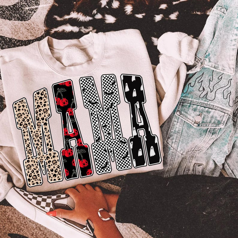 May include: Cream-colored sweatshirt with the word "MAMA" in a bold, patterned design. The letters feature leopard print, cherries, checkered patterns, and ghosts. A denim jacket and checkered slip-on shoe are also visible.