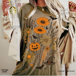 Peut inclure: T-shirt vert olive avec un motif floral sur le th&egrave;me d'Halloween. Le motif comprend des tournesols orange, une citrouille-lanterne, des chauves-souris, un fant&ocirc;me et un chapeau de sorci&egrave;re. Les mots "t&eacute;l&eacute;chargement num&eacute;rique" sont imprim&eacute;s en bas &agrave; droite.