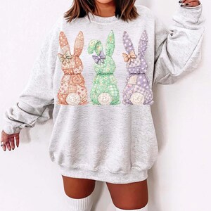 May include: A light gray sweatshirt featuring three colorful bunny designs. Each bunny is made of patchwork fabric in shades of orange, green, and purple, with floral and polka dot patterns. The bunnies have bows and fluffy white tails.