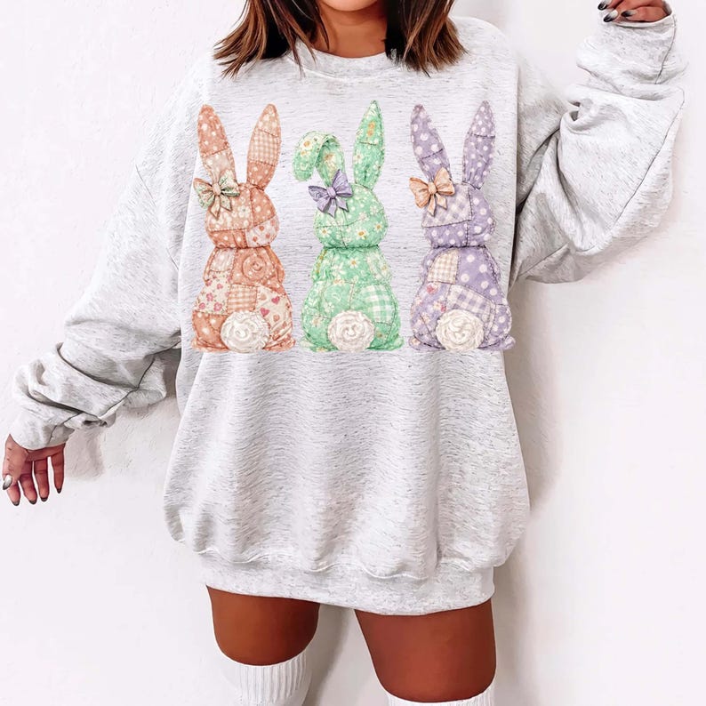 May include: A light gray sweatshirt featuring three colorful bunny designs. Each bunny is made of patchwork fabric in shades of orange, green, and purple, with floral and polka dot patterns. The bunnies have bows and fluffy white tails.