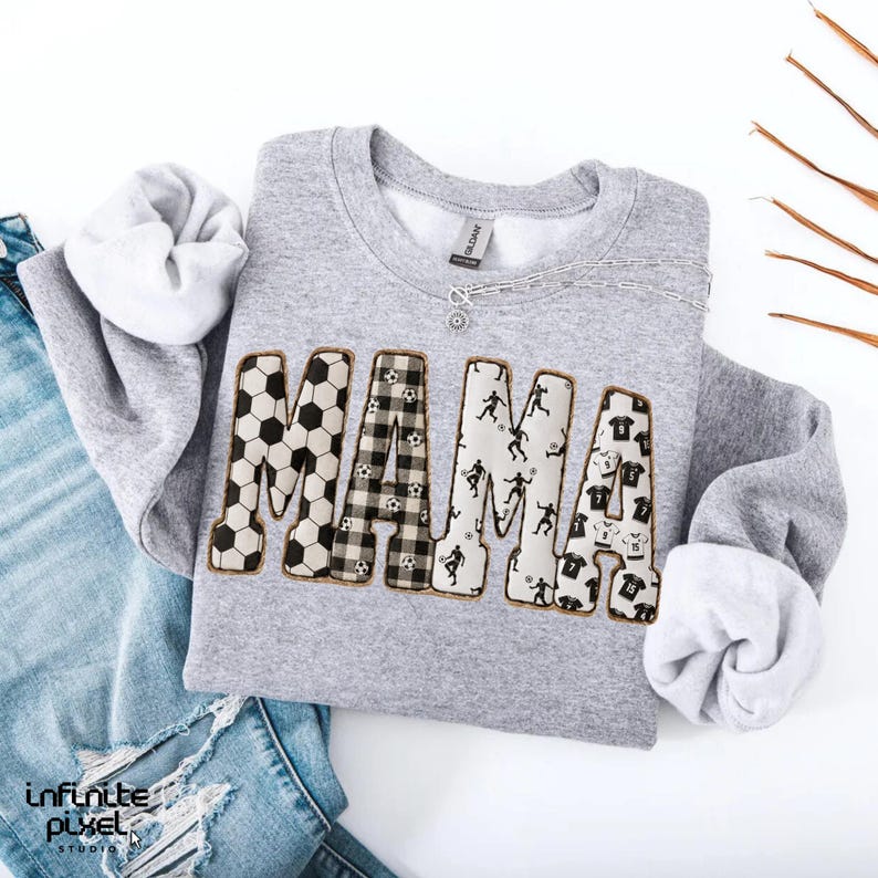 May include: A gray sweatshirt with the word "MAMA" in a soccer theme. The letters are decorated with soccer balls, plaid, soccer player silhouettes, and soccer jerseys. A silver necklace is draped across the sweatshirt.