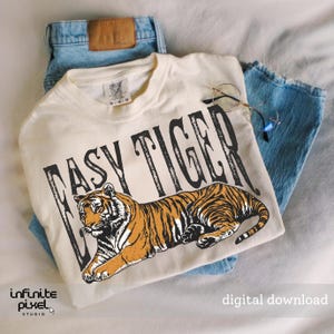 May include: A cream-coloured t-shirt featuring the words "EASY TIGER" above an orange and black tiger illustration. The t-shirt is placed on blue denim jeans, with round-framed glasses. The words "digital download" are also visible.
