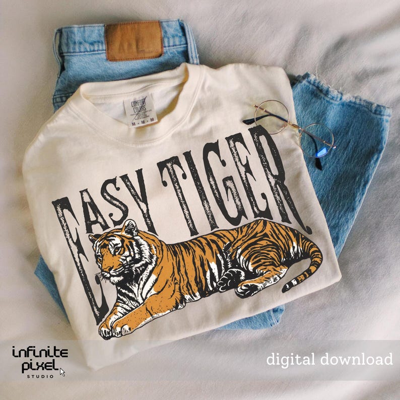 May include: A cream-coloured t-shirt featuring the words "EASY TIGER" above an orange and black tiger illustration. The t-shirt is placed on blue denim jeans, with round-framed glasses. The words "digital download" are also visible.