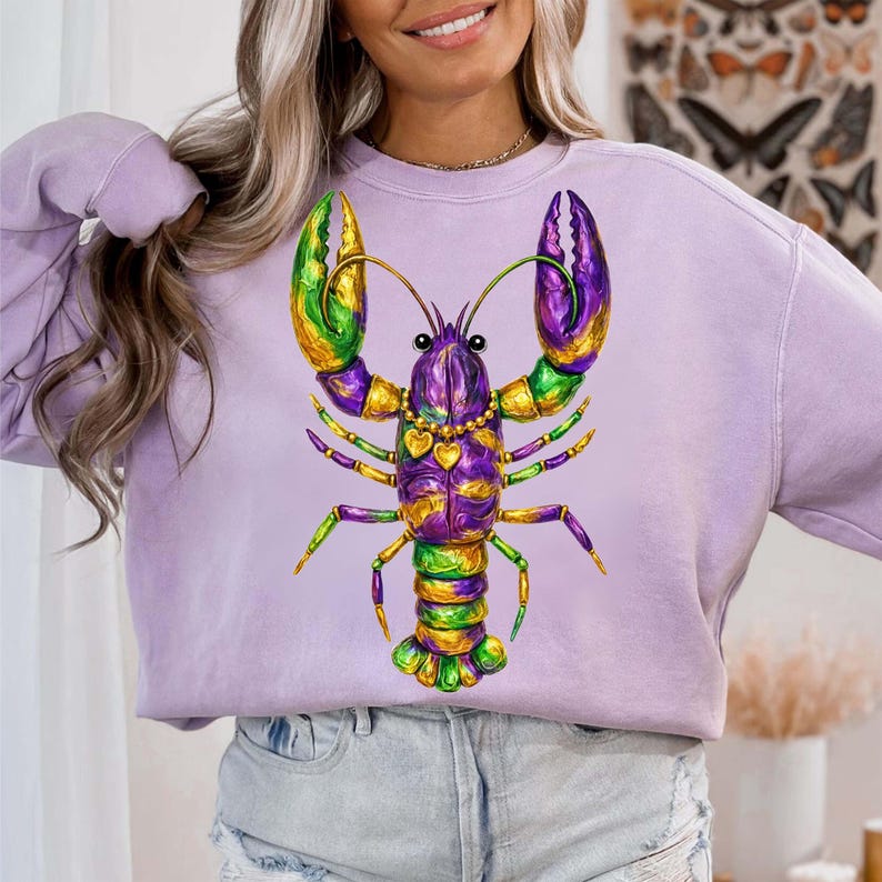 May include: A light purple sweatshirt featuring a colorful lobster design. The lobster is decorated with green, gold, and purple accents. The design includes a heart-shaped pendant. The sweatshirt is a comfortable, casual style.