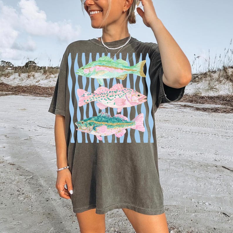 May include: Dark olive green t-shirt featuring a colorful fish design. The graphic includes three fish in shades of pink, green, and yellow, set against a blue striped background. The shirt has a relaxed fit.