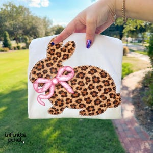 May include: White t-shirt with a leopard print bunny design and a pink bow. The bunny is outlined with a brown stitch. The image is held up by a person's hand. The text "infinite pixel studio" is in the bottom left corner.