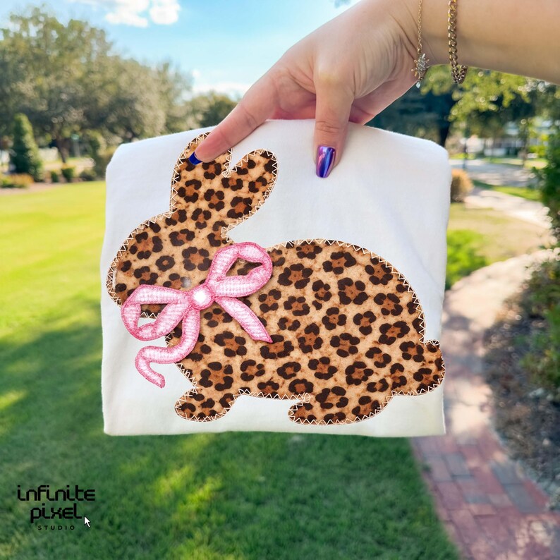 May include: White t-shirt with a leopard print bunny design and a pink bow. The bunny is outlined with a brown stitch. The image is held up by a person's hand. The text "infinite pixel studio" is in the bottom left corner.