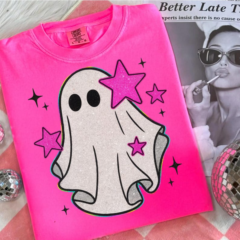 May include: A bright pink Comfort Colors sweatshirt featuring a white ghost graphic with black eyes and glittery pink stars. The design includes additional black star accents. The shirt is displayed on a pink and white checkered surface.
