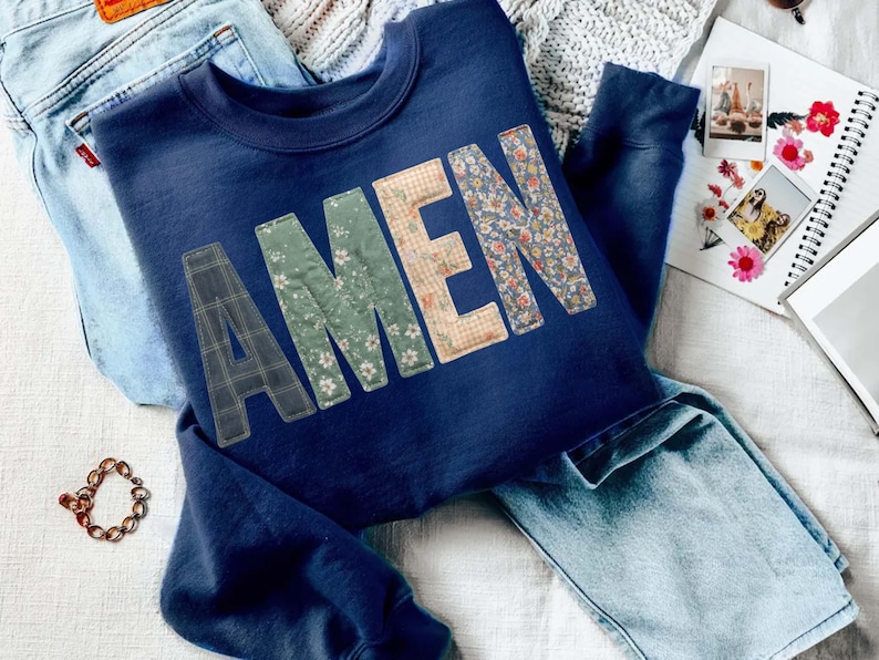 May include: A navy blue sweatshirt with the word "AMEN" appliqued across the front in various floral and plaid patterns. The sweatshirt is folded on top of a pair of light blue jeans. A gold chain bracelet and a notebook with photos are also visible.
