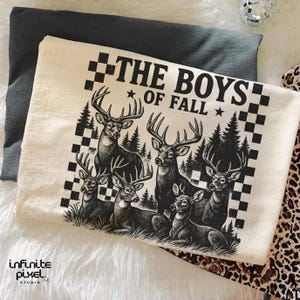 May include: Cream-colored t-shirt featuring a black and white graphic of six deer in a forest setting. The text "THE BOYS OF FALL" is printed above the deer. The shirt is folded on a gray surface.