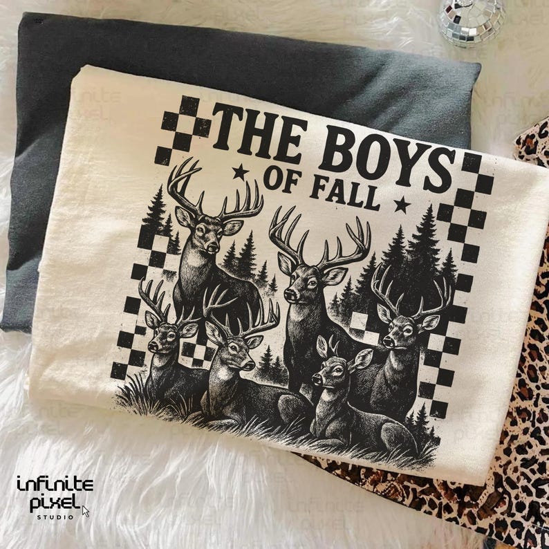 May include: Cream-colored t-shirt featuring a black and white graphic of six deer in a forest setting. The text "THE BOYS OF FALL" is printed above the deer. The shirt is folded on a gray surface.