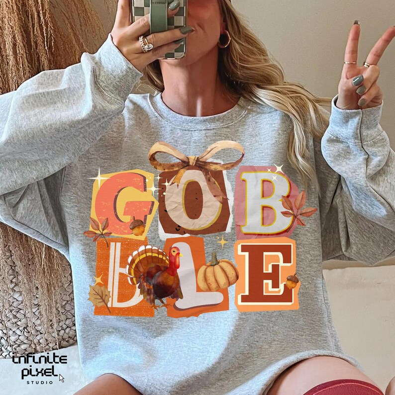 May include: A light gray sweatshirt with the word "GOBBLE" in colorful, block letters. The design includes a turkey, pumpkin, and fall leaves. The sweatshirt is a crew neck style.