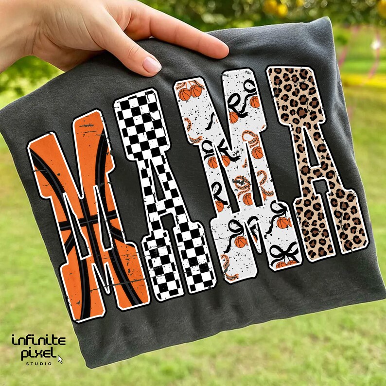 Puede incluir: Camiseta gris oscuro con la palabra "NANA" en letras grandes y estilizadas. Las letras presentan patrones de baloncesto, cuadros, baloncesto y estampado de leopardo. La camiseta est&aacute; sujeta por una mano sobre un fondo verde borroso.