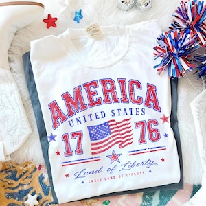 May include: White t-shirt with a distressed red and blue "AMERICA" graphic, the American flag, and the text "1776 Land of Liberty". The shirt is folded on top of a pair of gray pants, with patriotic decorations in the background.