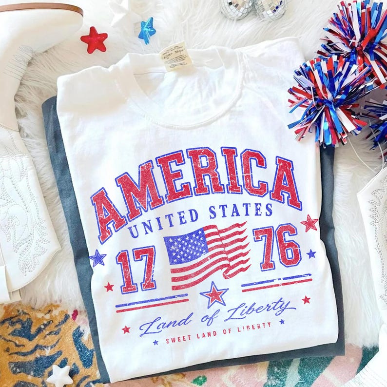 May include: White t-shirt with a distressed red and blue "AMERICA" graphic, the American flag, and the text "1776 Land of Liberty". The shirt is folded on top of a pair of gray pants, with patriotic decorations in the background.